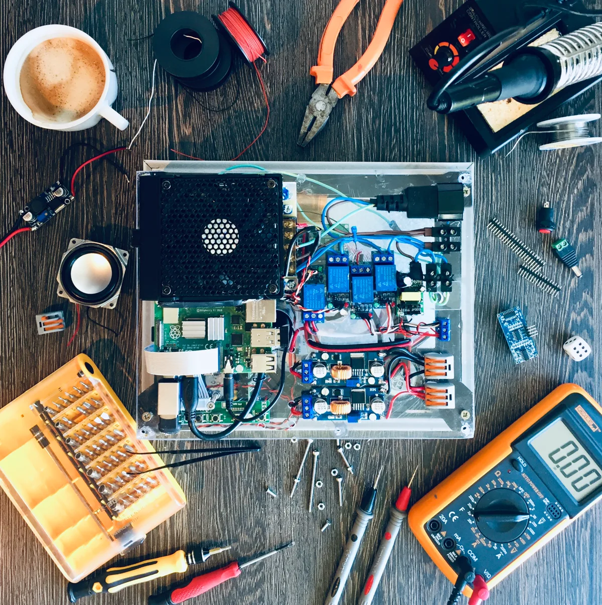 Overhead shot of a workbench: electronics, soldering iron, multimeter, coffee.